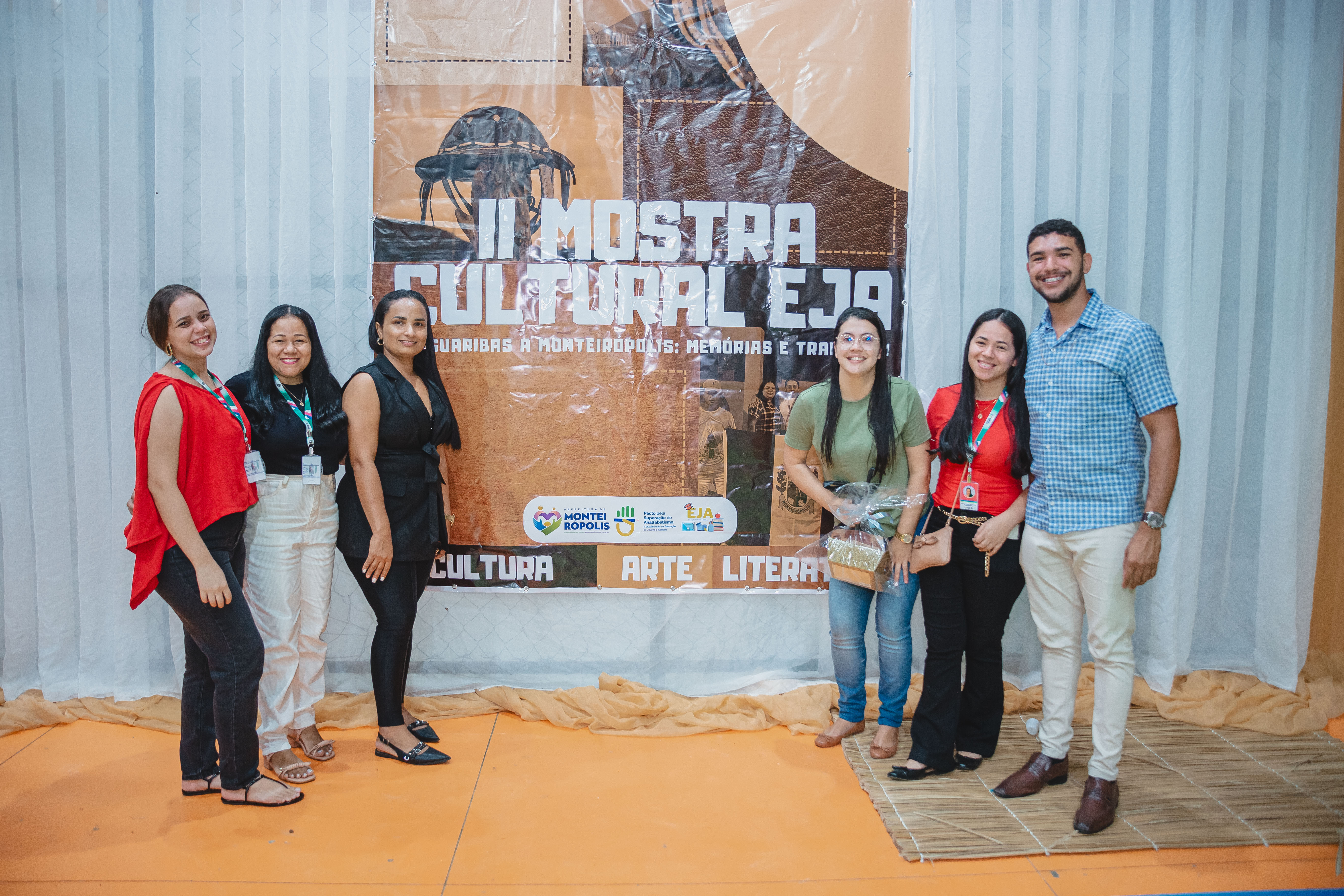 Pessoas posando para a foto em frente a cartaz que diz: "II Mostra Cultural EJA"
