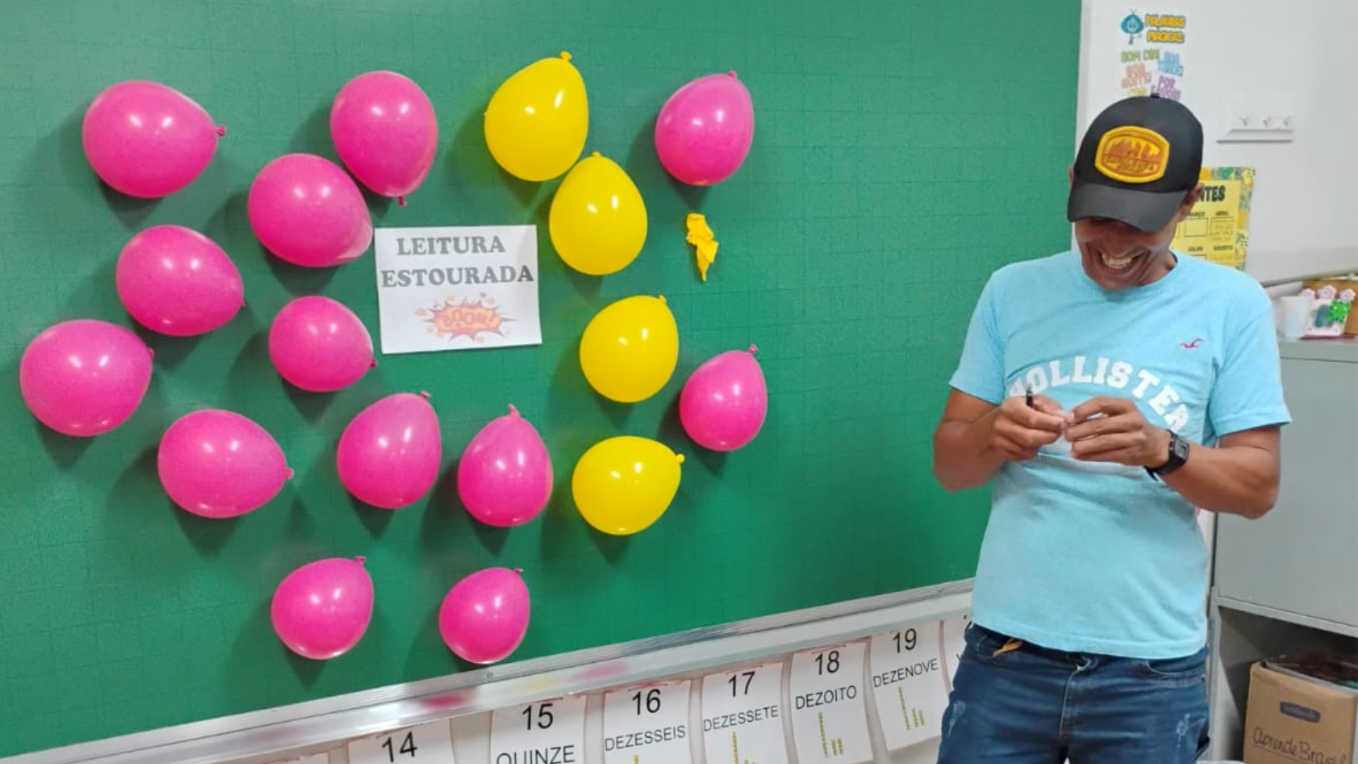 Homem sorri ao ler papel. Na lousa, há várias bexigas e um cartaz escrito "Leitura estourada"